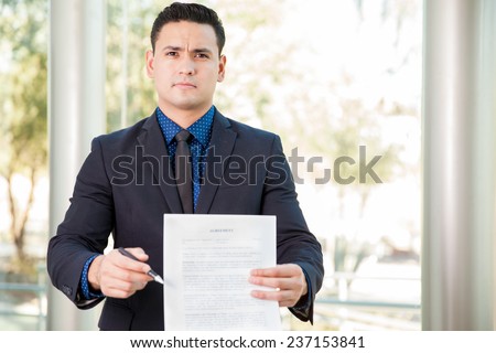 Portrait of a young Hispanic lawyer holding a contract and a pen and requesting a signature