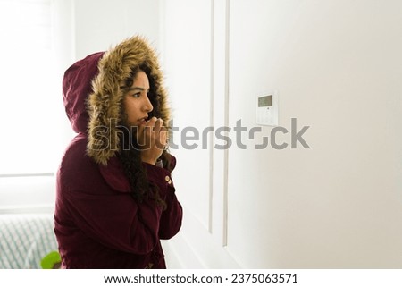 Sad hispanic woman wearing a winter jacket looking at the thermostat feeling very cold without the heating at home 商業照片 © 