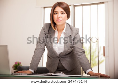 Successful and confident Latin business woman leaning on her desk and looking all serious