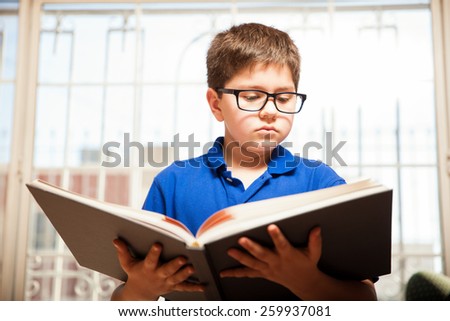 Young nerdy blond boy with glasses reading a big book at home