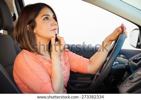 Young Latin woman putting some lipstick on and looking at herself in the mirror while driving a car