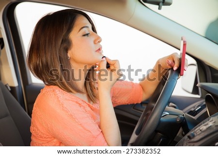 Profile view of a young brunette putting some lipstick on and looking at her cell phone while driving a car