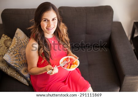 High angle view of a young pregnant woman sitting in the living room and eating some fruit from a bowl