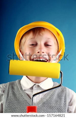 Beautiful cheerful blond boy wearing a yellow hard hat holding a yellow painting roller and laughing