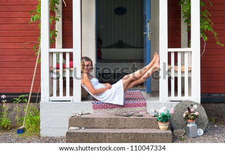 Attractive woman sitting on the steps of a veranda of a red, Swedish wooden house, relaxed summery atmosphere
