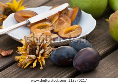 Stoned halved damson plums being prepared on a plate with whole purple plums in the foreground on a rustic wooden table