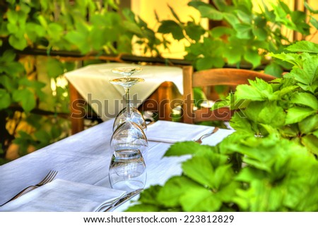 Crystal glasses are standing on a table with white table cloth in a restaurant framed with green plants