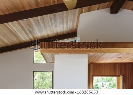 Detail of sloped wood beam ceiling with supports and wooden platform in house entryway. Trees can be seen outside the windows.