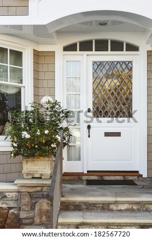 A white front door to a beige home, with an intricate, diagonal paned window. Also seen is a stone porch and stairs, railing, and a plant.