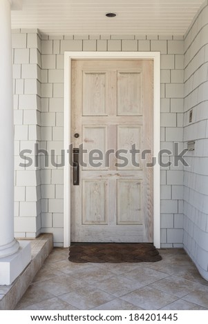 A wooden front door to a family home with raw, natural finish. House is white with shingles. Also seen is doormat, white column, and stone tile porch.