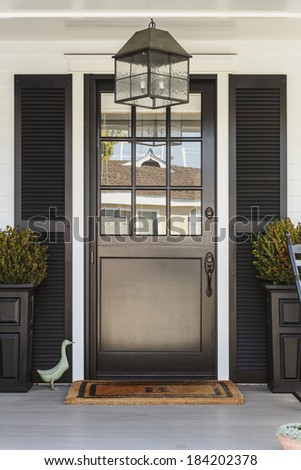 A black front door of a white family home with gray porch. The door is framed by black planters, black decorative shutters, and a light fixture. Also seen is a duck figurine, and a porch chair.