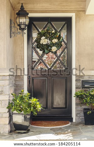 Black front door to home with flower wreath. A black front door to a home with an ornate flower wreath and classic light fixture. Also seen are door plants, and a stone porch.