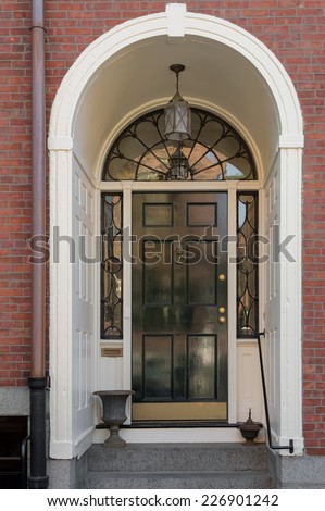 Ornate Entryway with Shiny Black Front Door, Overhead Lunette, and Hanging Lamp in White Archway of Brick Building