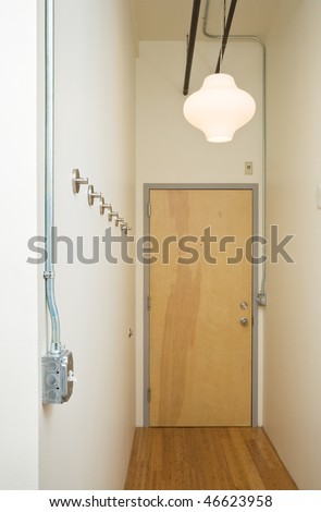 Looking down a sparse hallway towards a doorway. A light switch is exposed and coat racks line the wall. Vertical shot.