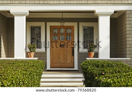 A concrete walkway bordered with hedged shrubs leads to the front door of a home. There are windows on either side of the door. Horizontal shot.