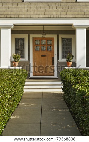 A concrete walkway bordered with hedged shrubs leads to the front door of a home. There are windows on either side of the door. Vertical shot.