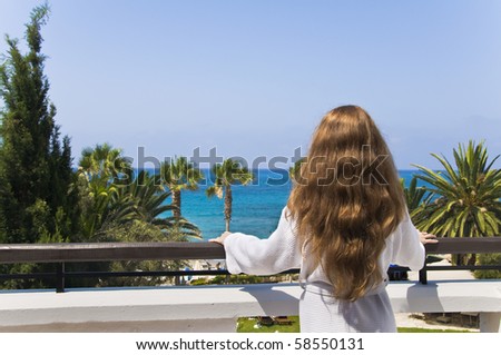 Young woman wearing white robe stands on the white hotel\'s balcony looks at the sea