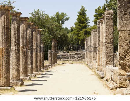 Two rows of old stone pillars stand on either side of a common area in the ruins of the ancient Greek city of Olympia.
