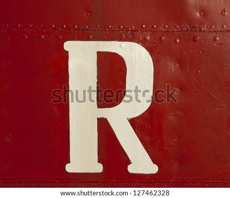 A big white letter \'R\', part of a ship\'s name, surrounded by a sea of intense red paint on the metal hull of the boat.