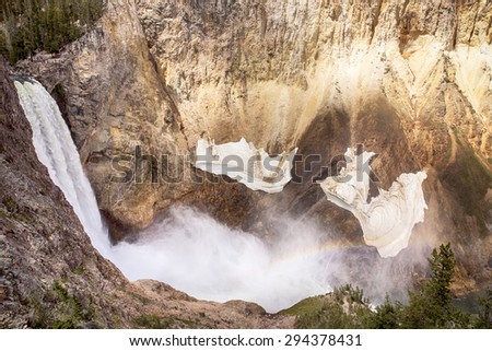 The waterfall at Lower Yellowstone Falls drops into the gorge of the Grand Canyon of Yellowstone National Park. A rainbow is visible in the mist and small snow fields hang on the canyon walls.