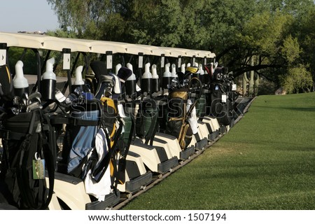 Golf carts are lined up at the starting area and are ready to start the day\'s round with a shotgun start tournament. The carts are loaded in the back with a variety of golf clubs and golf bags.