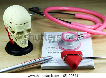 A doctors desk showing a green stethoscope and pen, resting on a sick certificate pad, with the other doctors tools of the trade on desk including a patella hammer and model of a human skull.