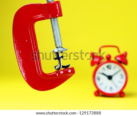 A silver pound symbol placed in a red clamp with a yellow background, with a red alarm clock in the background indicating the pressure on the pound sterling.
