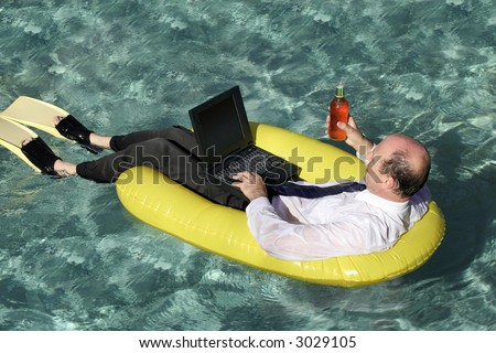Businessman in suit, tie with a laptop in one hand and a beer in another, in an inflatable boat on the ocean wearing flippers, surrounded by the clear blue ocean