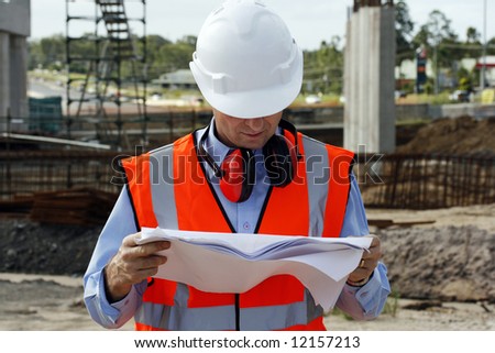 Engineer looking thoughtful, examining the plans, wearing a white safety helmet, high visibility orange vest and wearing ear muffs around his neck at the work site.
