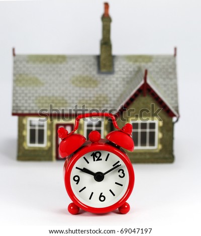 A red Alarm clock in front of a house , asking the question is it time to sell or buy.