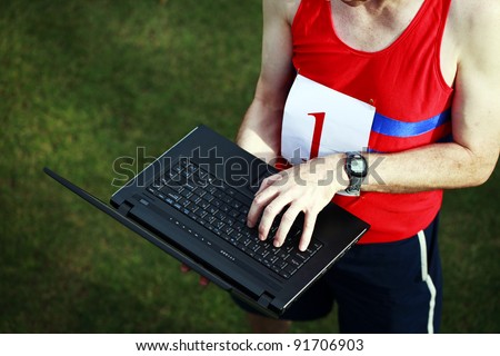 A close up of a businessman dressed in running attire with the number one on his chest, working on his laptop outside.