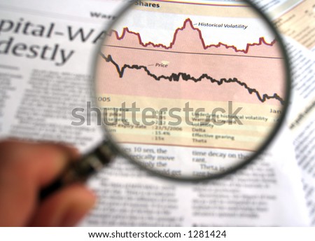 Hand holding a magnifying glass focusing on a chart in the business section of the newspaper.