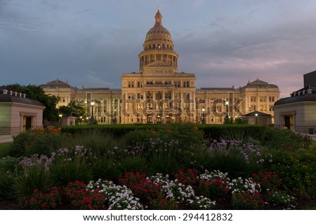The Capitol Building in Austin, Texas with colorful flower beds in the foreground, lit up by street lights as the sun sets and a cloudy sky grows dark as dusk.