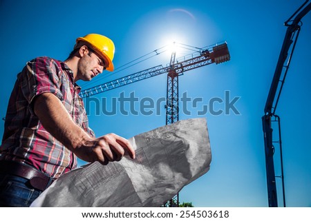 Real Engineer looking at his blueprint plan on construction site, crane and concrete pump seen in the background