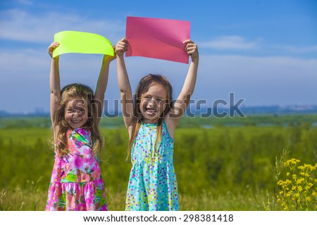Two baby girls in summer dresses on the nature of holding the colored sheets of paper. Summer, sun, green, meadow.
