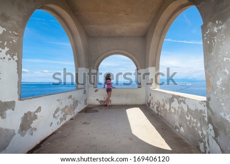 Young woman in the pink shirt standing next to the arch shaped stone window and looking at the sea.