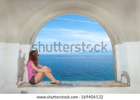 Young woman in the pink shirt sitting on the arch shaped stone window and looking at the sea.