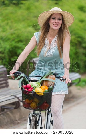 Attractive blonde woman with straw hat riding a bike with basket full of groceries.