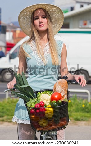 Attractive blonde woman with straw hat posing next to bike with basket full of groceries.