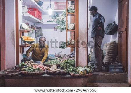 VARANASI, INDIA - 19 FEBRUARY 2015: Grocer sitting on ground with customer standing in doorway small vegetable shop on street market. Post-processed with grain, texture and colour effect.