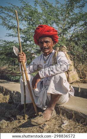 GODWAR REGION, INDIA - 14 FEBRUARY 2015: Elderly Rabari tribesman with red turban sits and holds ax and stick. Post-processed with grain, texture and colour effect.