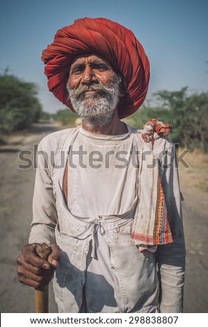 GODWAR REGION, INDIA - 14 FEBRUARY 2015: Elderly Rabari tribesman with big red turban and cane stands on road. Post-processed with grain, texture and colour effect.