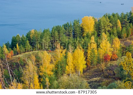 Excellent autumn landscape with yellow, red and green leaves on the trees. The tree line down to the blue river. In the river, the fishermen are fishing from boats.