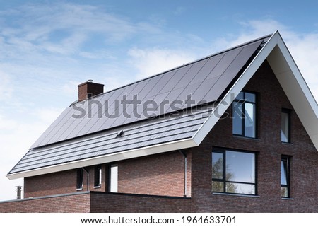 Solar panels mounted on the roof of a modern new-build house in a street in Heerenveen, Friesland, the Netherlands with sun and blue cloudy sky. Sustainable energy