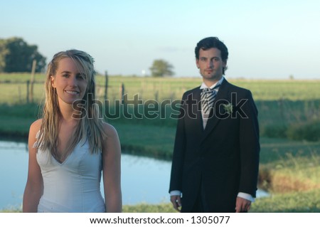A bride and groom with the groom in the background. Lake-side country scene at sunset.