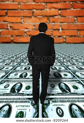 businessman standing on a floor of dollar banknotes with a brick wall in front of him