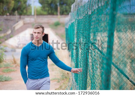 A Good looking male model walking past a fence, wearing trousers and a long sleeved t-shirt