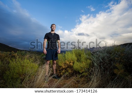Male fitness model running allong a trail in the field, wearing a black shirt and shorts, with big clouds ooverhead in the sky.