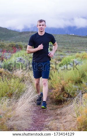 Male fitness model running allong a trail in the field, wearing a black shirt and shorts, with big clouds ooverhead in the sky.