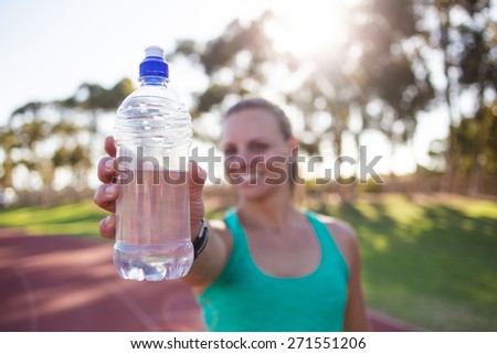 Close up wide angle view of a female fitness athlete holding up a bottle of water with a smile on her face. Back light natural light.